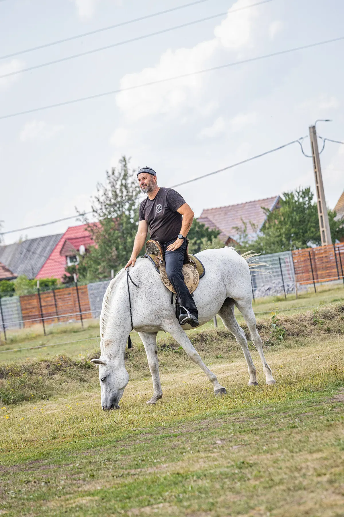 Un călăreț stă relaxat pe un cal alb care paște într-o pajiște de la marginea satului.