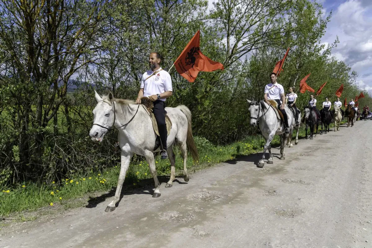 O procesiune de călăreți cu steaguri roșii înaintează pe un drum de țară.