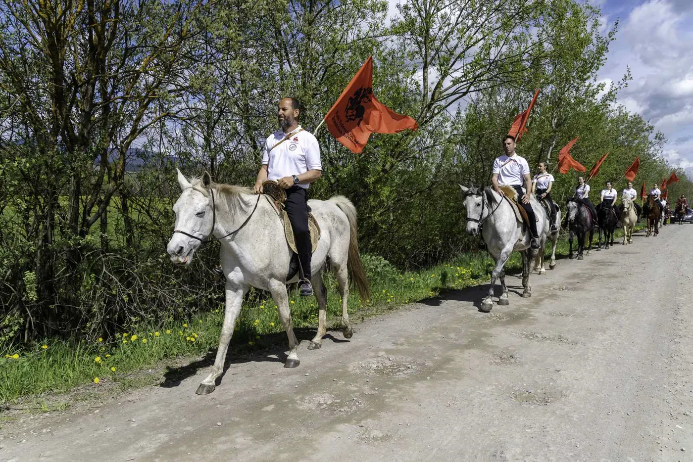 O procesiune de călăreți cu steaguri roșii înaintează pe un drum de țară.